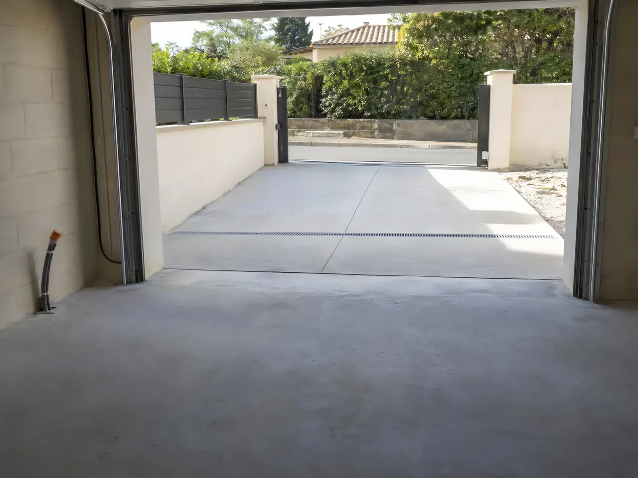 Intérieur d'un garage neuf vide : sol en béton, grande porte ouverte sur une allée bétonnée et un portail