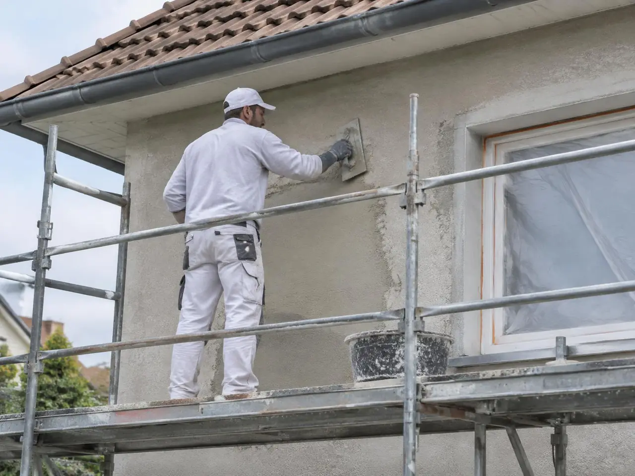 Artisan sur l'échafaudage qui rebouche à la taloche des zones de crépi gris, fenêtre protégée par un film