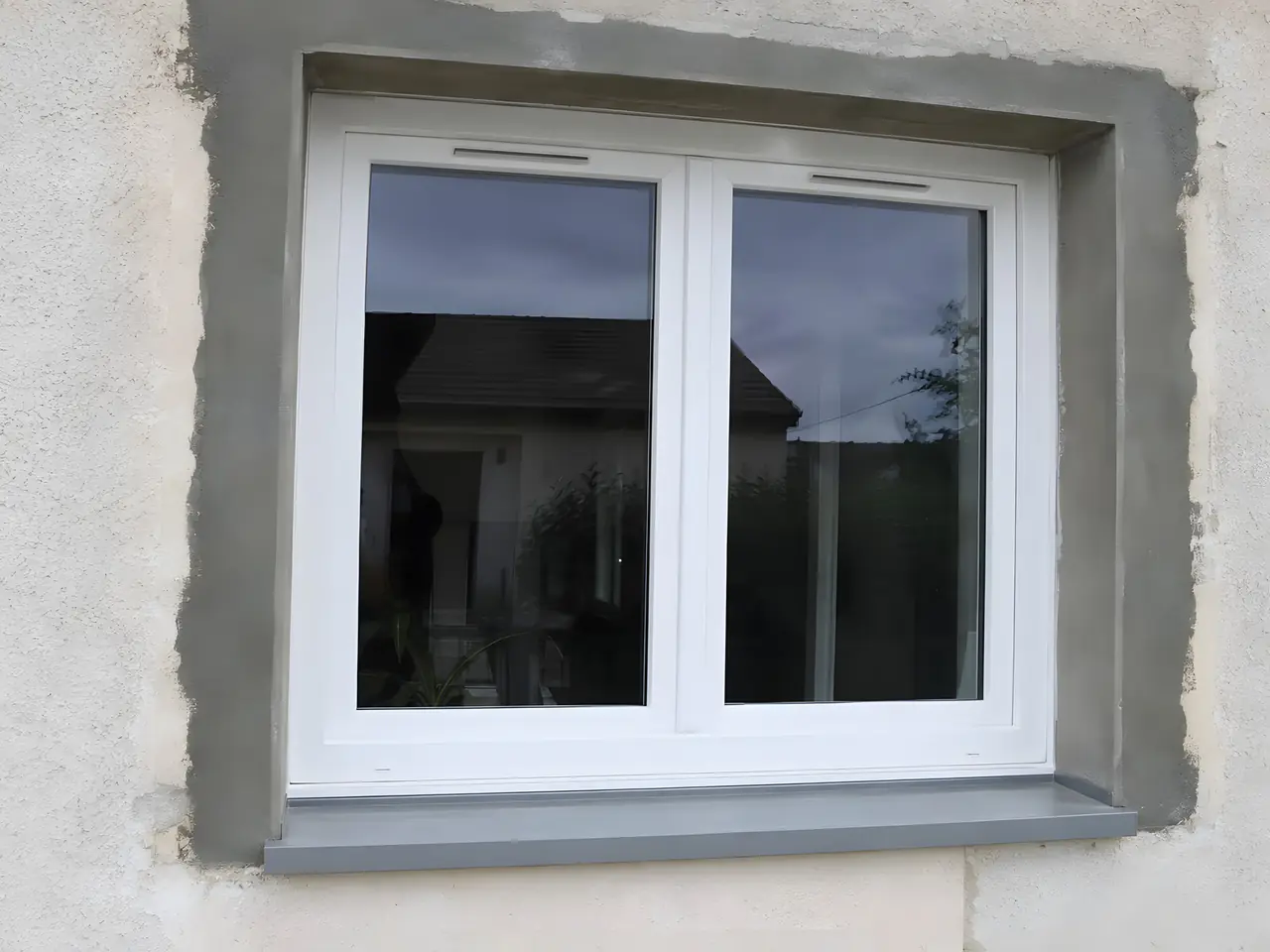 Fenêtre blanche neuve dans une façade enduite, tableaux encore en enduit gris brut et appui gris