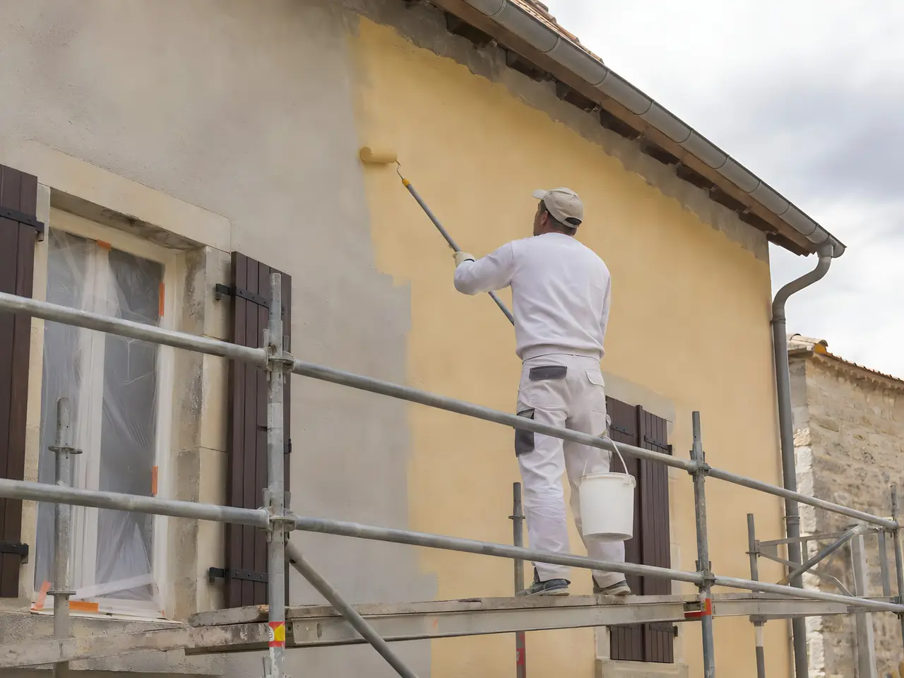 Peintre sur un échafaudage qui passe au rouleau une peinture jaune sur une façade grise aux volets bruns