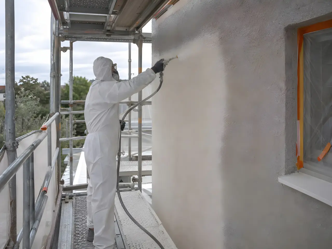 Artisan en combinaison et masque qui projette un enduit à la machine sur un mur, depuis un échafaudage