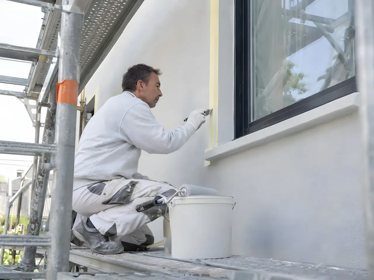 Peintre accroupi sur un échafaudage qui peint au pinceau le bas d'un encadrement de fenêtre, pot blanc à côté