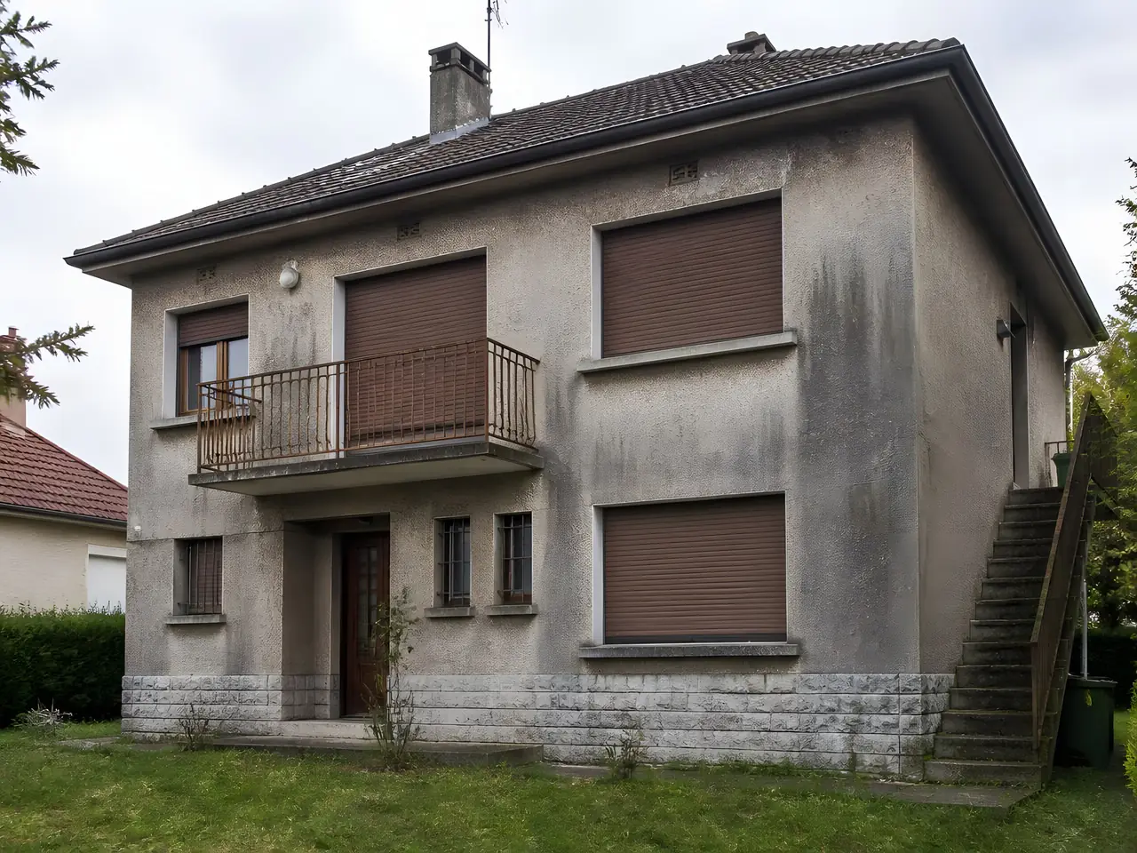 Pavillon à étage au crépi gris noirci par les années, coulures sombres sous les fenêtres, volets roulants bruns