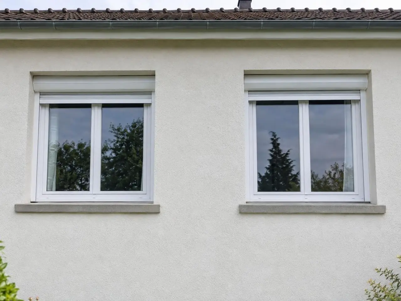 Façade en crépi blanc cassé propre et uniforme sous deux fenêtres blanches à volet roulant et leurs appuis gris clair