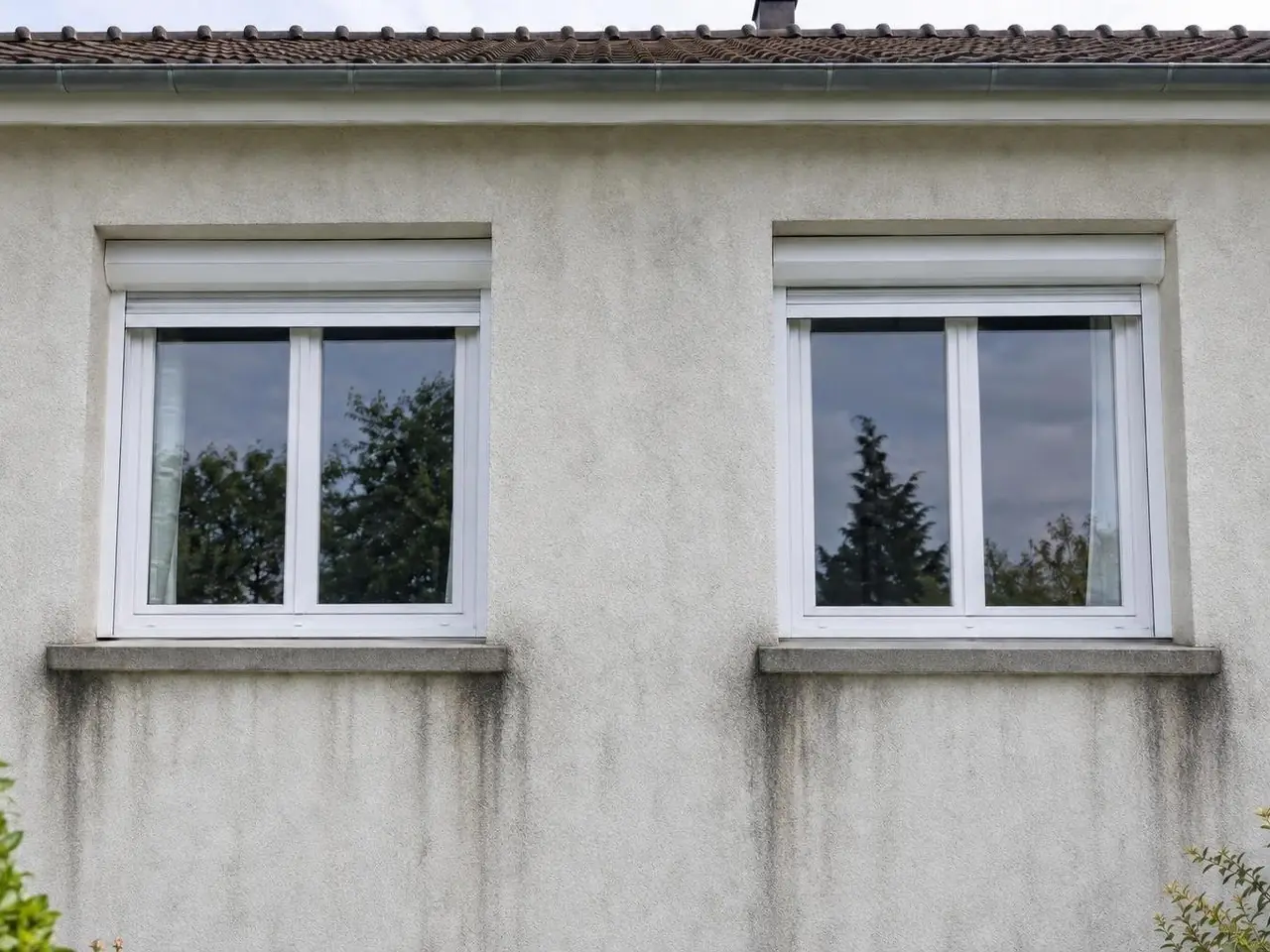 Façade en crépi blanc cassé grisé, longues traînées noires sous les appuis de deux fenêtres blanches à volet roulant