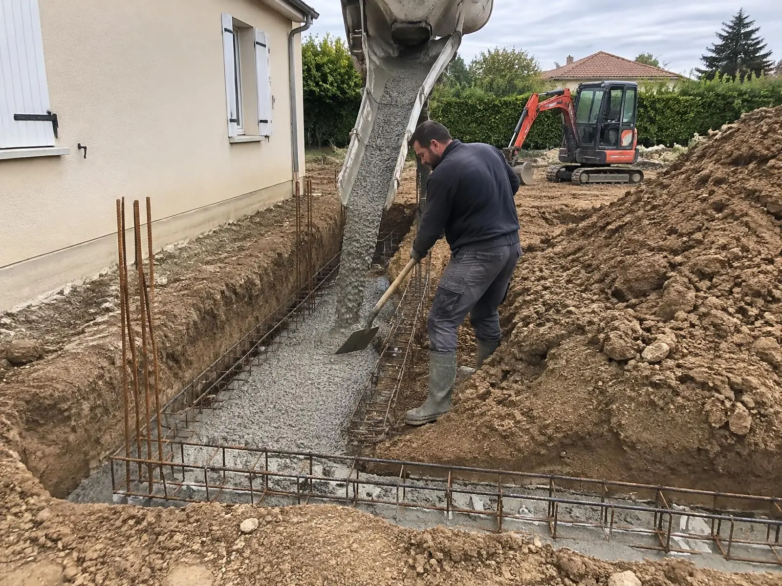 Maçon qui étale à la pelle le béton coulé dans les fondations ferraillées d'une extension, contre un pavillon.