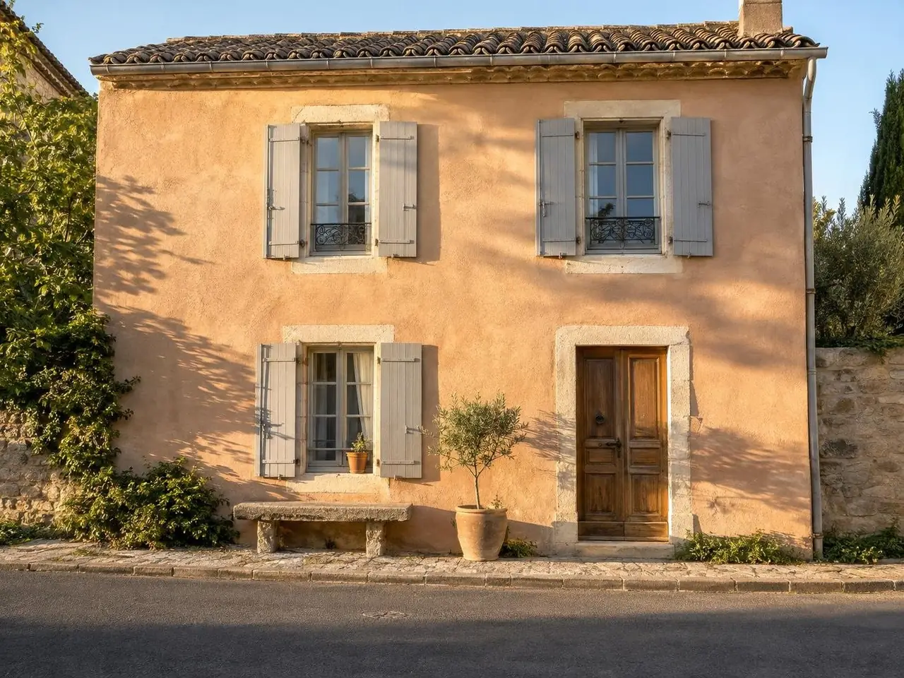 Façade de maison de village peinte à la chaux ton sable rosé, encadrements en pierre claire, volets gris, banc de pierre