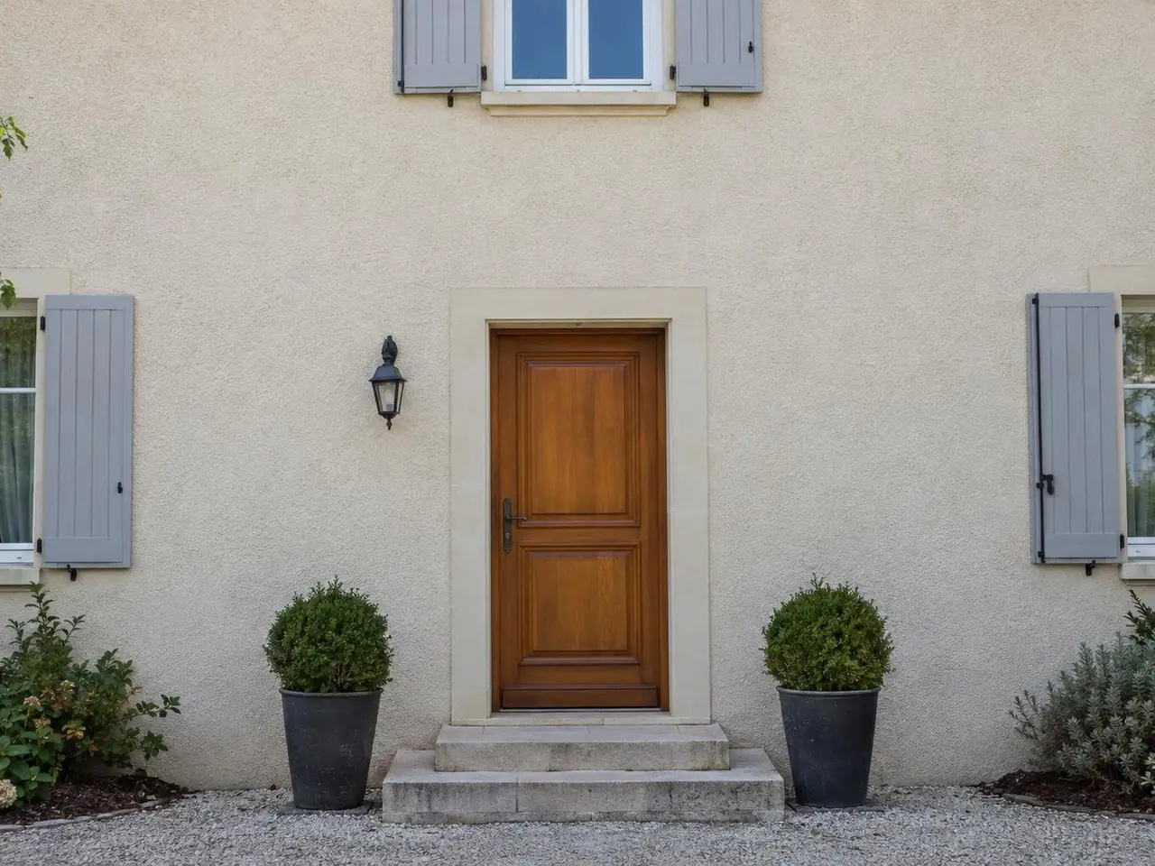 Façade en crépi beige uniforme au-dessus d'une porte d'entrée en bois verni, applique noire et deux buis en pot