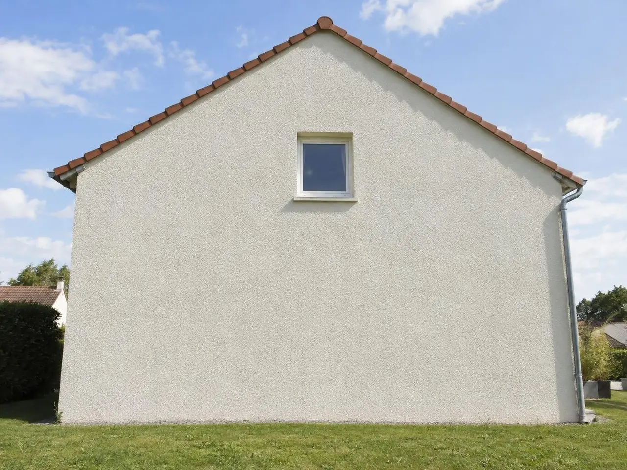 Pignon de pavillon en crépi gratté blanc cassé neuf et uniforme sous un toit de tuiles rouges, pelouse au pied du mur