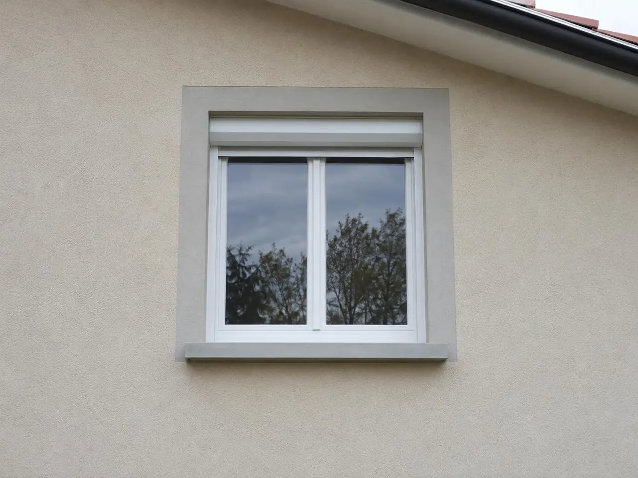 Fenêtre blanche à volet roulant encadrée d'une bande d'enduit gris clair, appui en béton lisse aux arêtes nettes