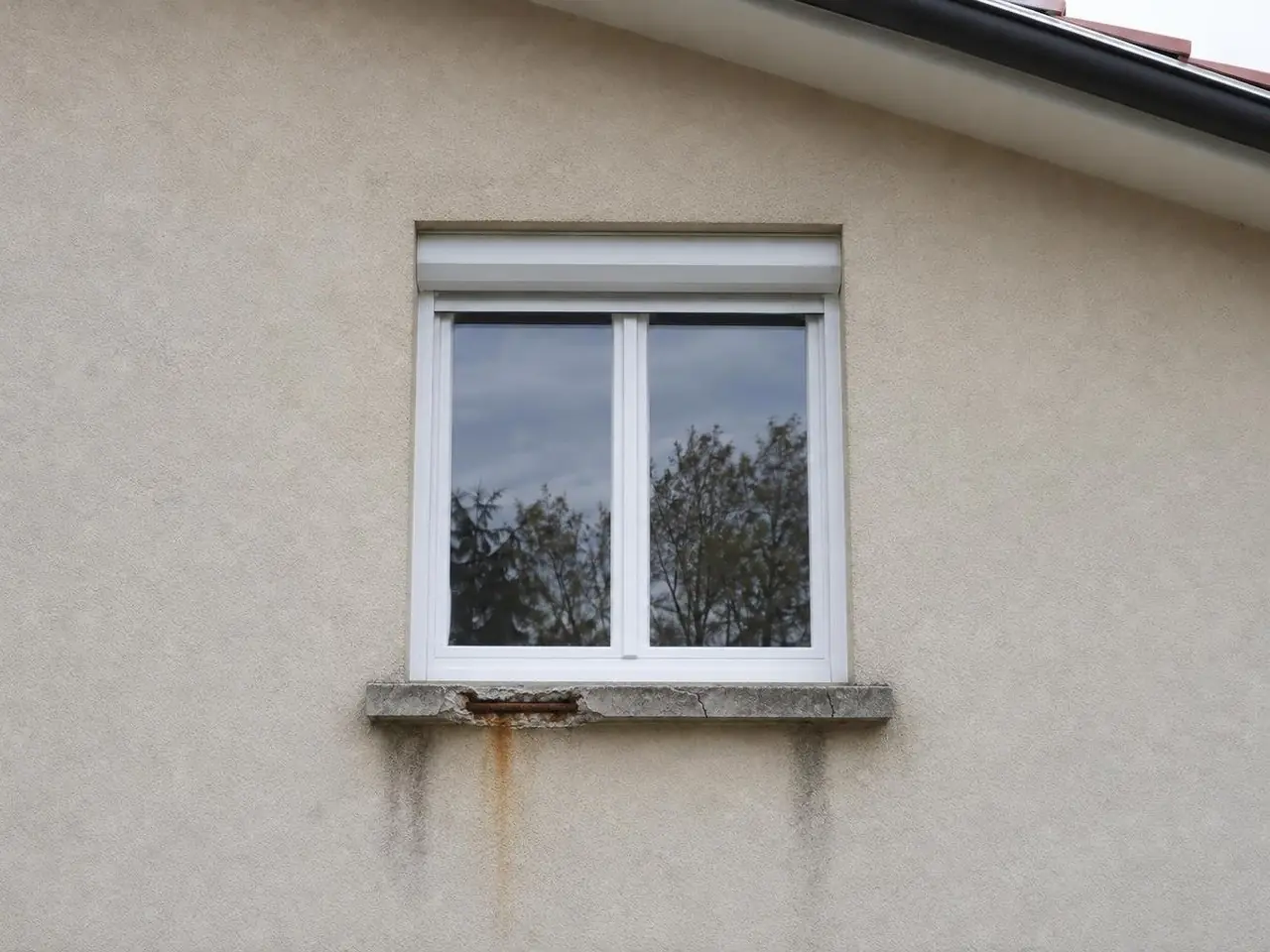 Fenêtre blanche à volet roulant au-dessus d'un appui en béton éclaté, ferraille rouillée et coulures brunes sur le crépi
