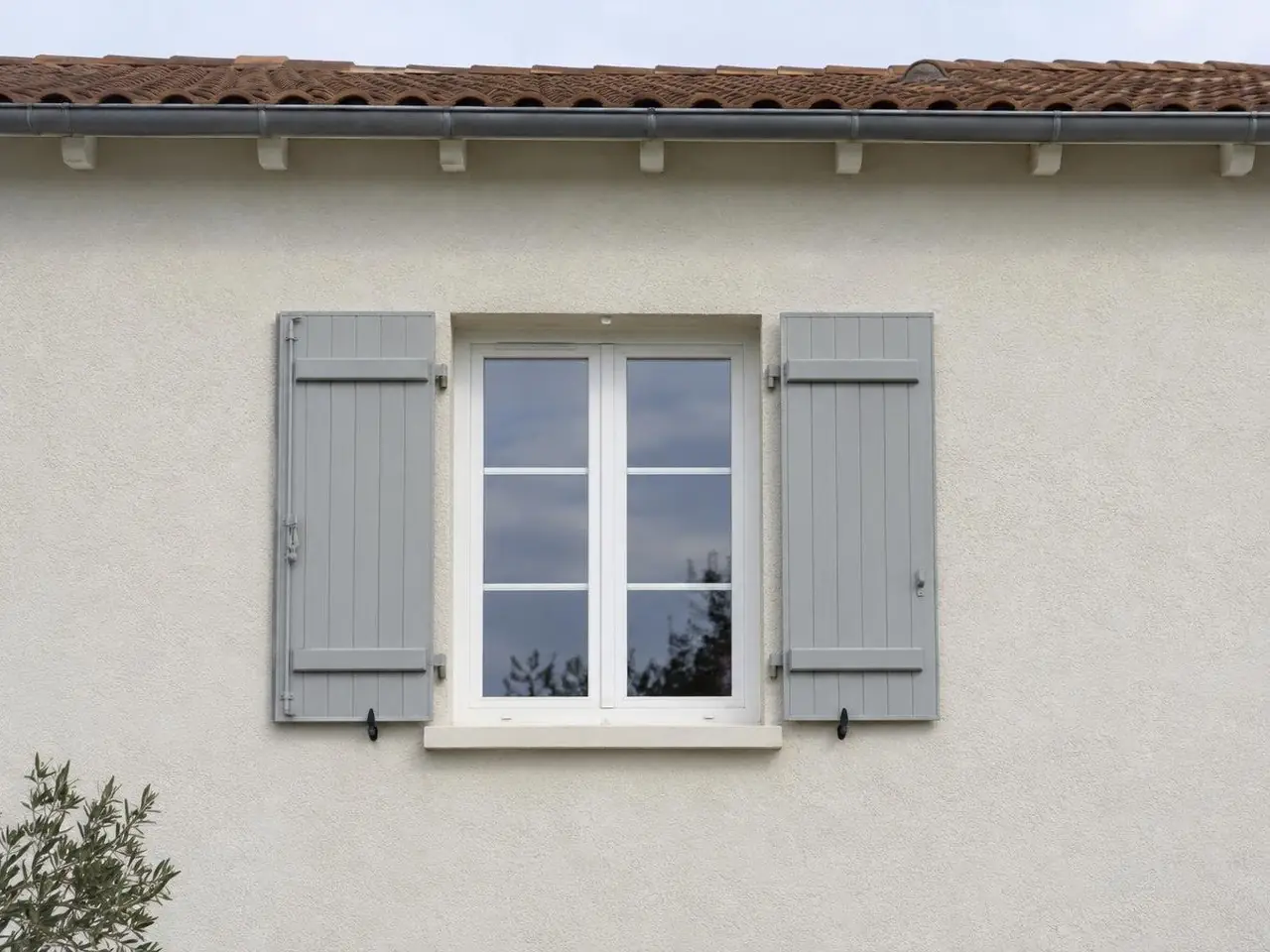 Fenêtre blanche à volets gris clair dans un crépi blanc cassé uniforme, aucune fissure entre la fenêtre et le toit