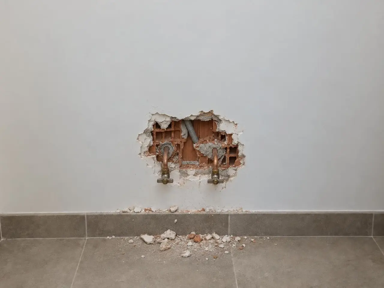 Mur blanc de salle de bains éventré autour de deux tuyaux en cuivre, briques creuses à nu, débris de plâtre au sol