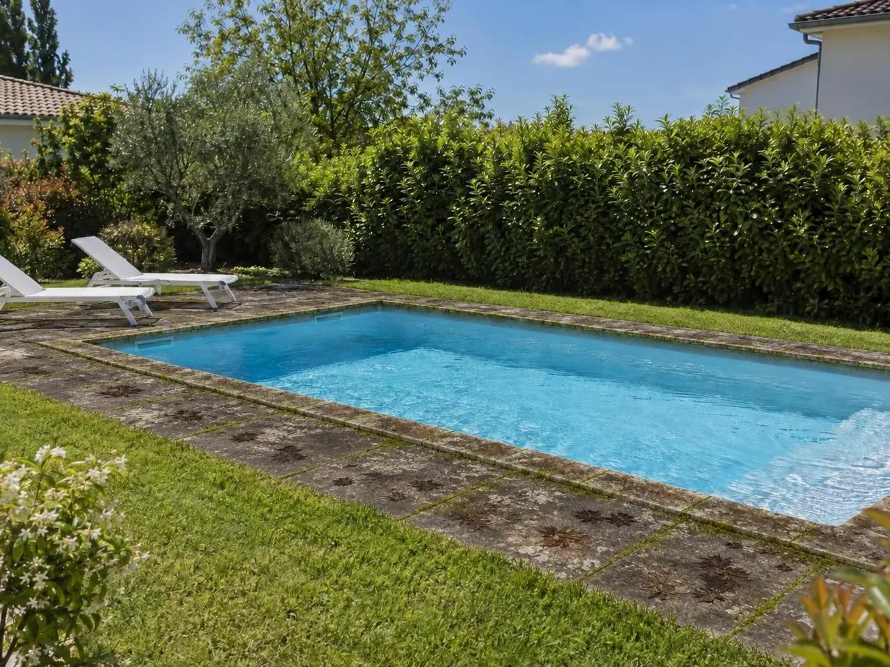 Tour de piscine en dalles grisâtres tachées de vert et d'auréoles de feuilles, bains de soleil devant une haie